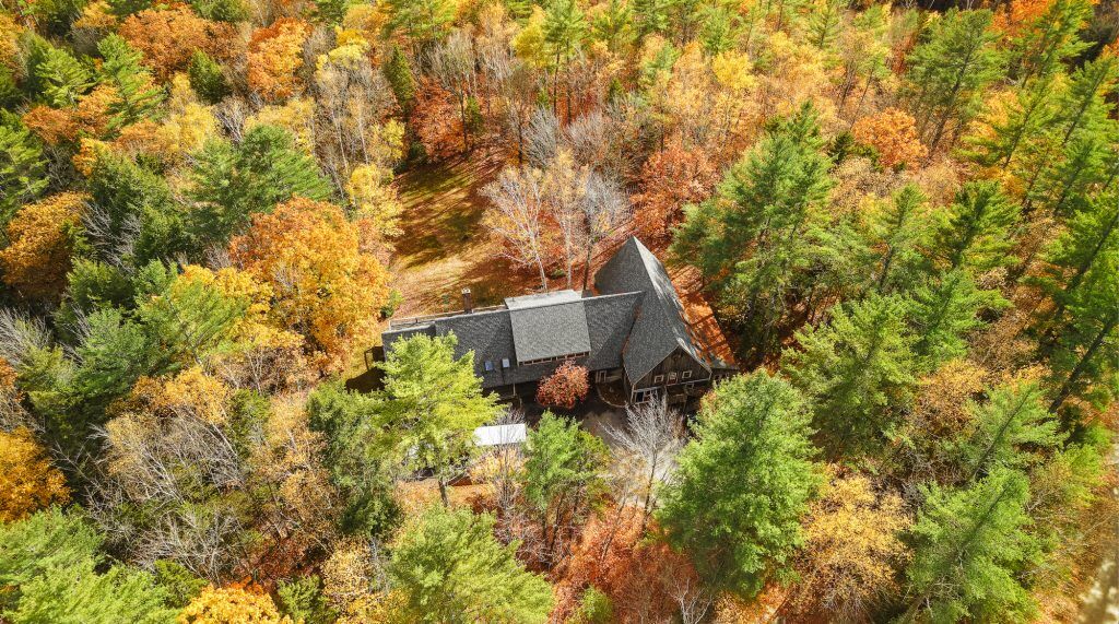 The image shows a house surrounded by a vibrant forest during the fall season. The trees display a mix of green, yellow, and orange foliage, creating a colorful canopy. The house has a dark roof and appears to be nestled within the trees, offering a sense of seclusion and natural beauty.